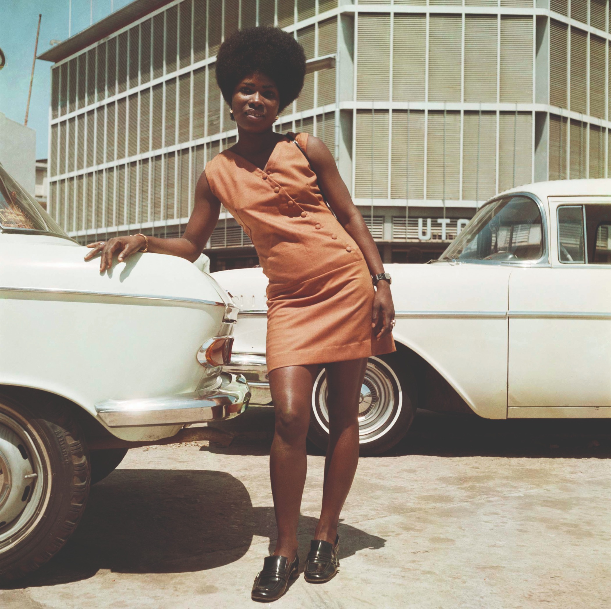 Woman posing outside the United Trading Company headquarters, Accra, Ghana (1971). © James Barnor. Courtesy of Galerie Clémentine de la Féronnière A woman wearing an orange mini dress leans on a white car as she poses outside the United Trading Company headquarters in Accra, Ghana, 1971